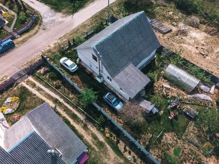 Aerial photo of a rural house with garden, greenhouse, and parked cars in Belarus. Shot in clear daylight.