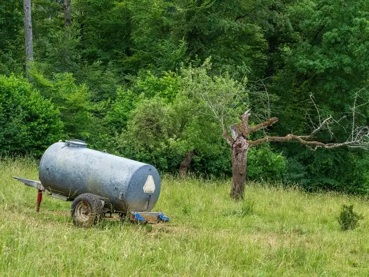An old water tank sits in a lush meadow surrounded by dense green foliage and trees.
